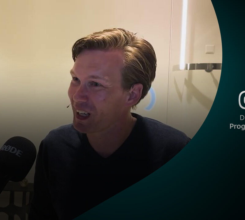 Geert Jan Hahn in gesprek met microfoon voor zich, met bloemen op de achtergrond.