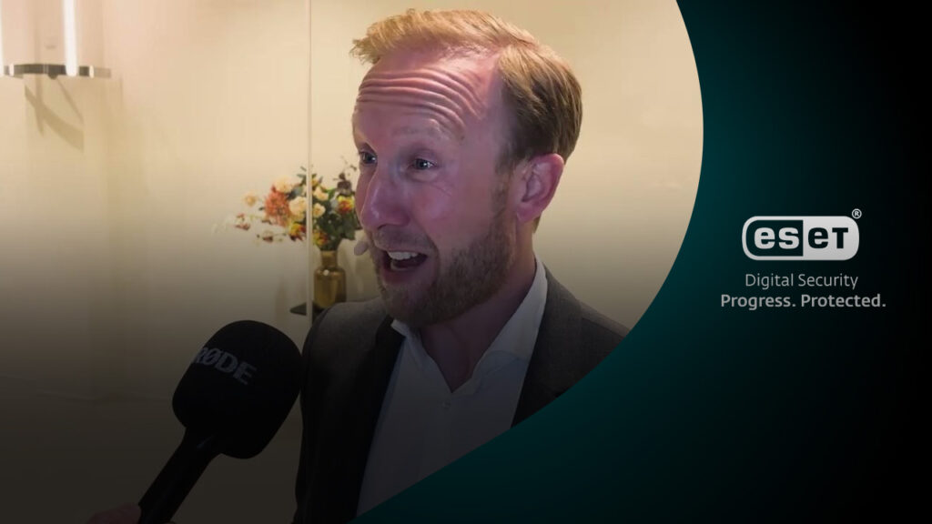 Raymond Mens reageert tijdens interview met microfoon, bloemen op de achtergrond.