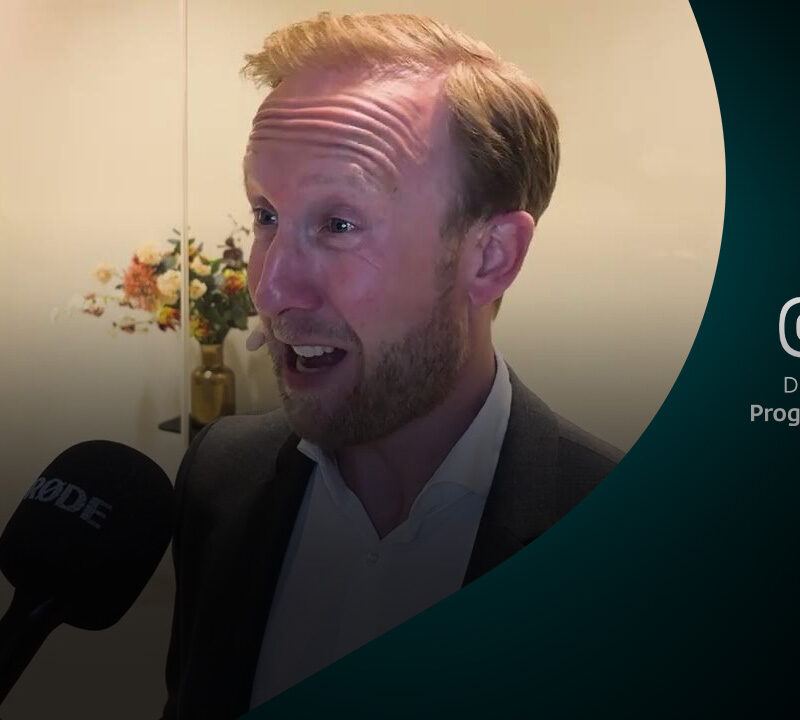 Raymond Mens reageert tijdens interview met microfoon, bloemen op de achtergrond.